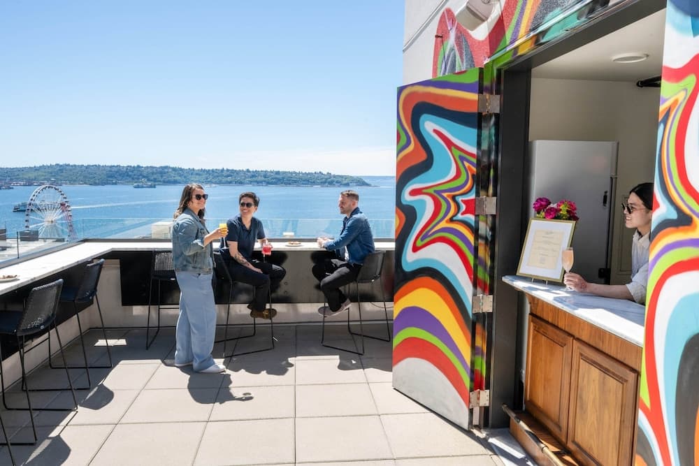 A vibrant rooftop bar with colorful abstract art walls offers a lively social scene where guests enjoy drinks under sunny skies, overlooking a serene waterfront with a distant Ferris wheel, perfect for urban travelers seeking unique city views.