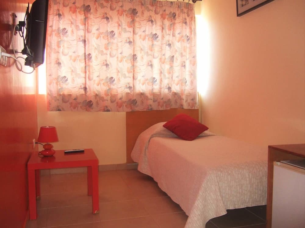 A cozy hotel room features a neatly made bed with a red pillow, a vibrant red side table with a lamp, floral curtains, and warm lighting, ideal for travelers seeking comfort and simplicity.