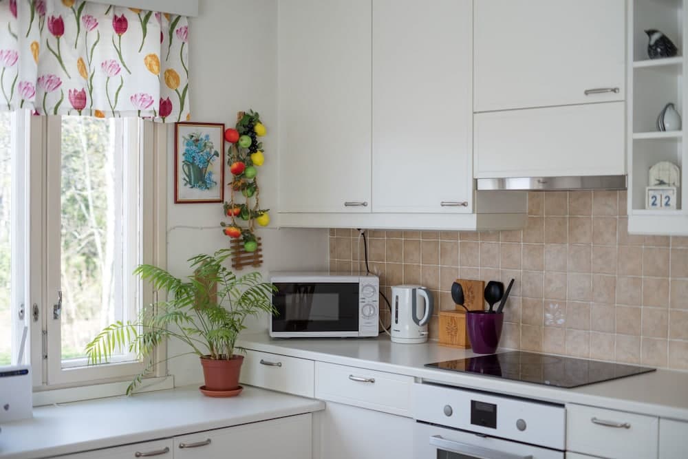 A cozy kitchen with white cabinetry, a tiled backsplash, and a window adorned with floral curtains, featuring a microwave, kettle, and vibrant indoor plant, ideal for travelers seeking a homey cooking experience.