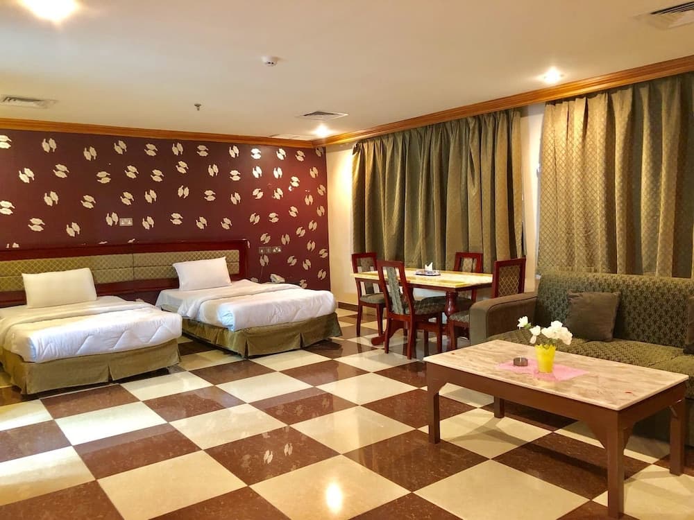 A cozy hotel room features twin beds against a patterned wall, a checkered floor, a dining table with chairs, and a sofa by the curtains, ideal for travelers seeking comfort and functionality.