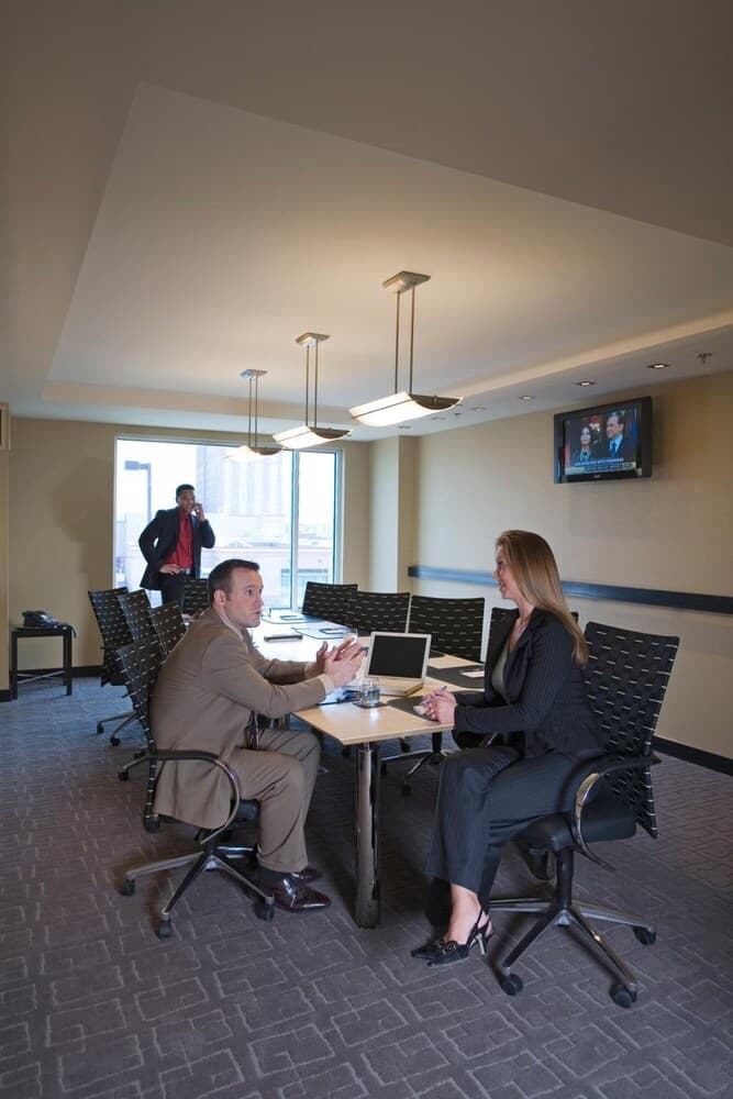 A professional meeting unfolds in a modern conference room with sleek furniture, a mounted TV displaying news, and large windows letting in natural light, ideal for business travelers seeking a productive workspace.