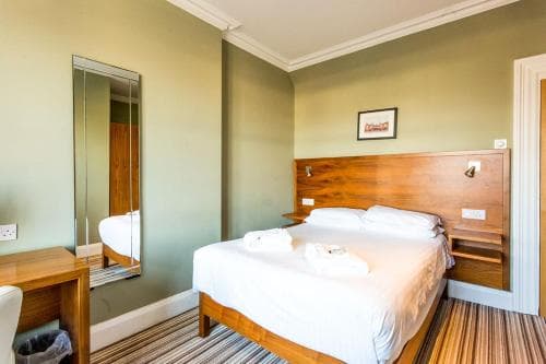 A cozy hotel room with soft green walls, a neatly made bed with white linens, wooden furnishings, and a mirror reflecting the space, ideal for travelers seeking comfort and simplicity.