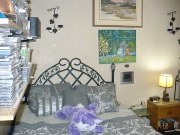 A cozy bedroom scene features a metal bedframe adorned with plush pillows and a purple teddy bear, set against a wall decorated with framed art and a warm bedside lamp, ideal for travelers seeking a comfortable and inviting accommodation.