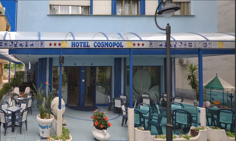 A vibrant blue Hotel Cosmopol with outdoor seating and potted plants invites travelers to relax in its cheerful, seaside atmosphere, perfect for a leisurely vacation getaway.