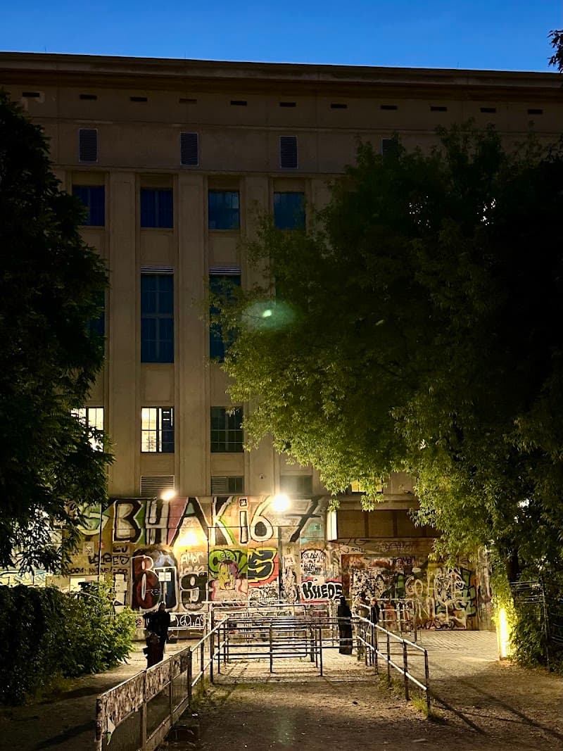 A nighttime view of an industrial building with vibrant graffiti under the glow of streetlights, surrounded by lush trees, evokes a mysterious urban atmosphere ideal for nightlife exploration in Berlin.