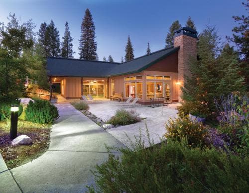 A cozy cabin nestled among tall pines at dusk, featuring a warmly lit patio and lush landscaping, evokes a serene retreat ideal for nature lovers seeking a peaceful getaway.