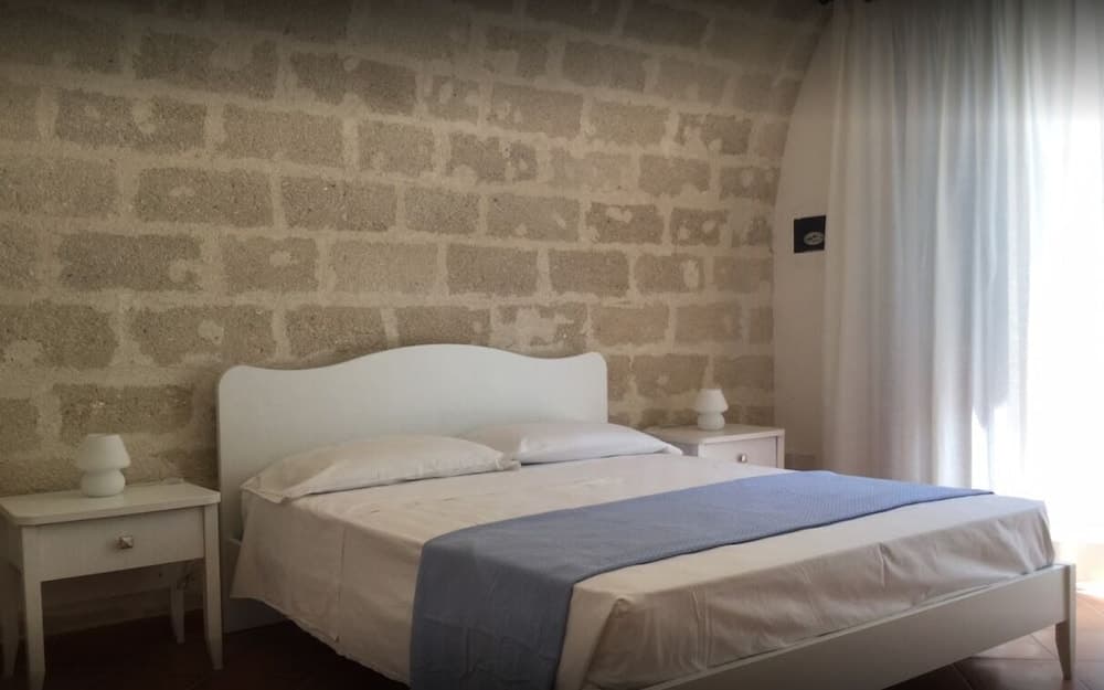 A cozy bedroom with a rustic stone wall, white furnishings, and soft natural light through sheer curtains offers a serene retreat for travelers seeking a tranquil stay.