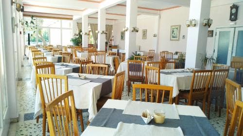A spacious, warmly lit indoor dining area with wooden chairs, white tablecloths, and decorative plants creates an inviting atmosphere for travelers seeking a cozy restaurant experience.