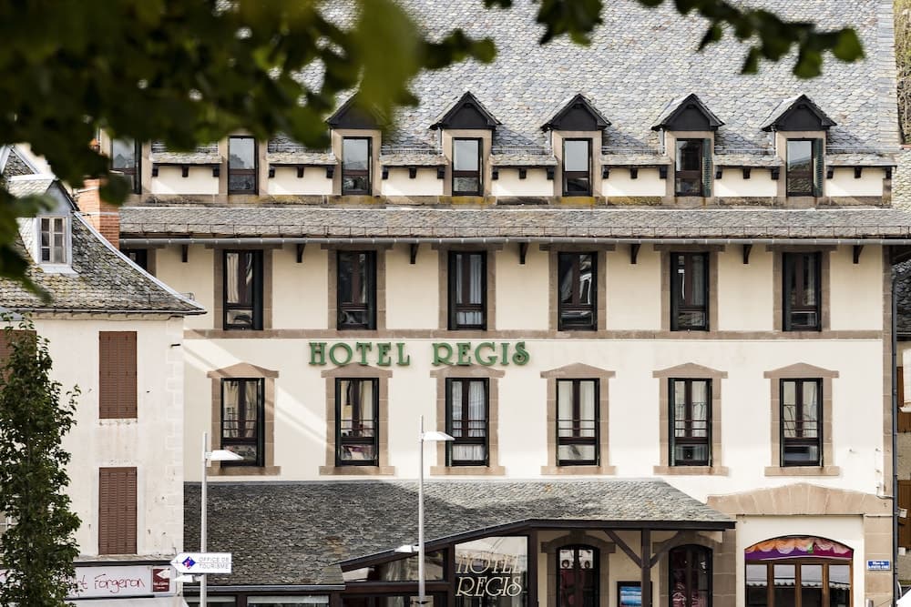 A charming European hotel with a slate roof and green signage framed by lush foliage offers travelers a cozy retreat in a picturesque town setting.
