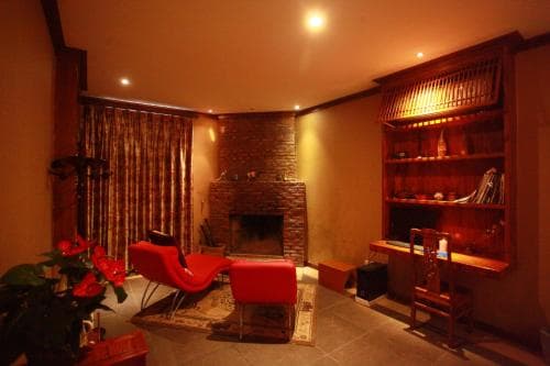 A cozy indoor lounge features warm lighting, a brick fireplace, red seating, and wooden shelves filled with books and decor, creating an inviting atmosphere ideal for relaxation or a quiet getaway.