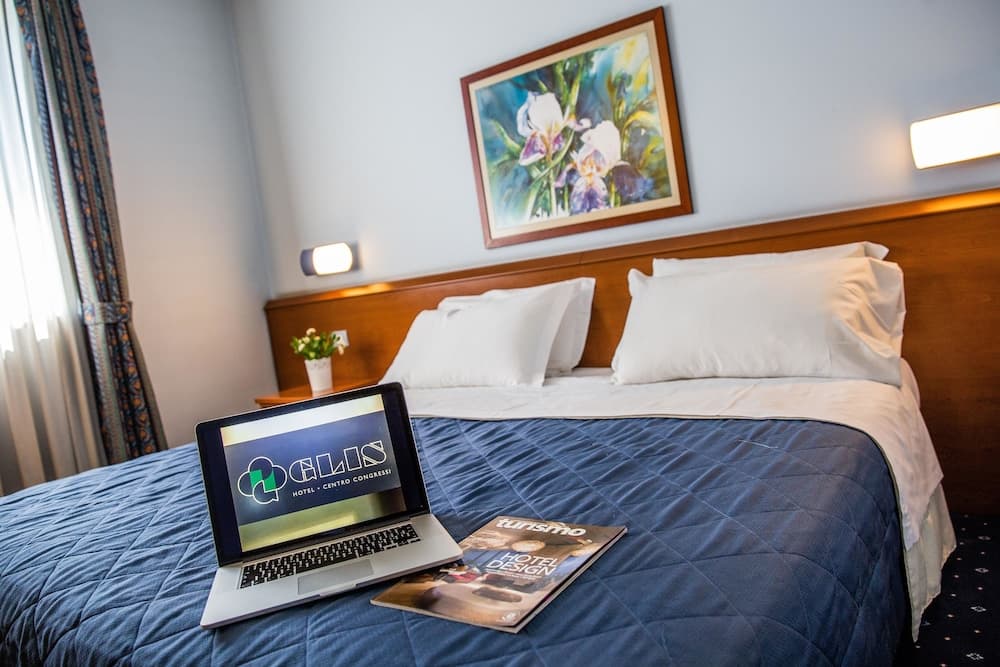 A cozy hotel room features a neatly made bed with a laptop displaying "GLIS Hotel - Centro Congressi" and a magazine on it, set against soft lighting and floral artwork, ideal for business travelers seeking comfort and productivity.