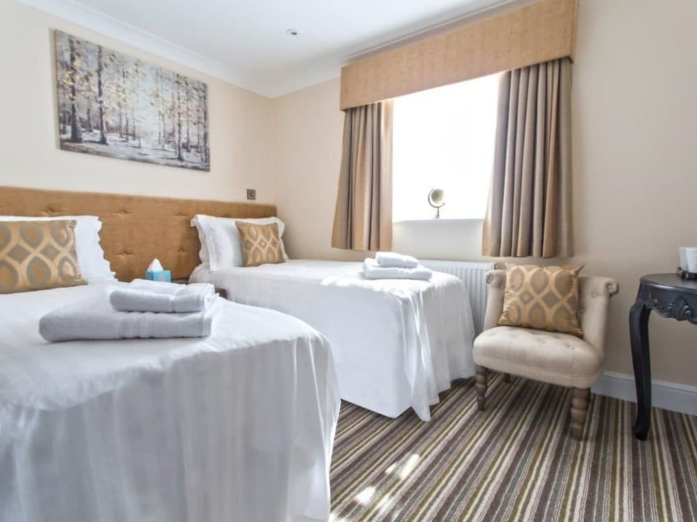 A cozy hotel room features two neatly made beds with white linens, a plush armchair, and a window with beige curtains, creating a serene atmosphere ideal for travelers seeking comfort and relaxation.