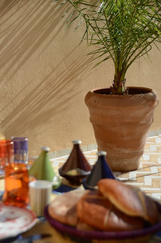 A serene outdoor dining setup features a terracotta-potted palm casting shadows on a beige wall, surrounded by colorful tagines, fresh bread, and drinks, ideal for a relaxing culinary adventure in a Mediterranean-style courtyard.