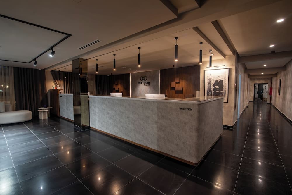 A modern hotel lobby exudes sophistication with sleek black tiles, a minimalist reception desk, hanging pendant lights, and a framed portrait, ideal for travelers seeking upscale accommodations.