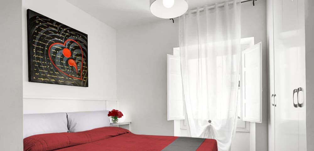A cozy bedroom with white walls, a red bedspread, and a heart-themed artwork creates an inviting atmosphere, perfect for travelers seeking a romantic getaway.