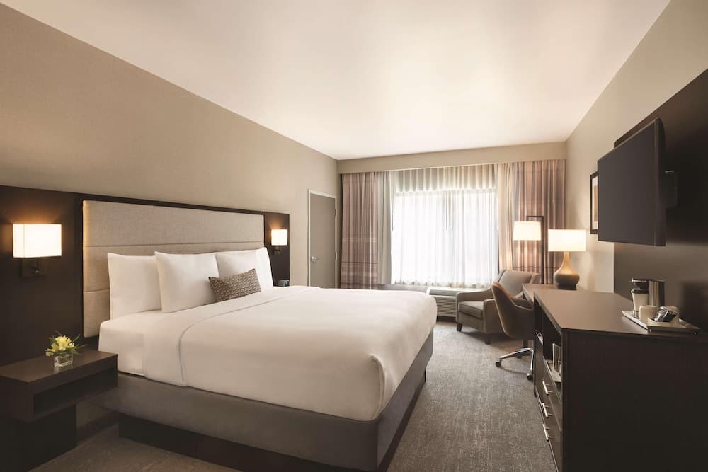 A cozy hotel room features a neatly made bed, soft lighting, a comfortable seating area by the window, and modern amenities, ideal for travelers seeking relaxation and convenience.