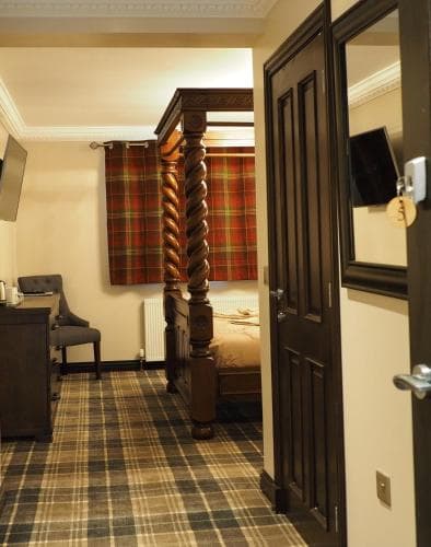 A cozy hotel room features a traditional four-poster bed, tartan-patterned carpet and curtains, creating a warm and inviting atmosphere ideal for travelers seeking comfort and cultural charm.