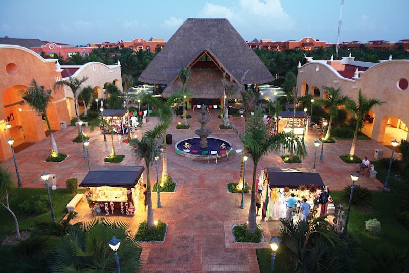 A vibrant evening scene at an all-inclusive resort in Mexico, featuring a central courtyard with a thatched-roof structure, palm trees, a fountain, and illuminated walkways, ideal for travelers seeking a luxurious tropical getaway.