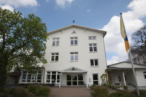 A serene white building with a welcoming entrance and colorful flag stands amidst lush greenery under a bright blue sky, ideal for travelers seeking a peaceful accommodation in a quaint town setting.