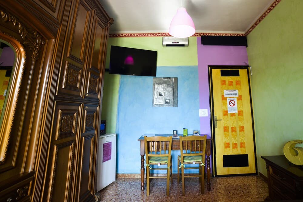 A cozy, colorful hotel room features a small dining table with chairs, a vibrant wall mural, a mounted TV, and ornate wooden furniture, ideal for travelers seeking a charming and unique accommodation experience.