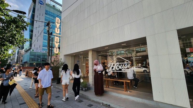 A bustling urban street in Seoul features the modern facade of NYU NYU House with its inviting storefront, where pedestrians stroll past, creating a lively and trendy atmosphere ideal for city exploration and shopping.