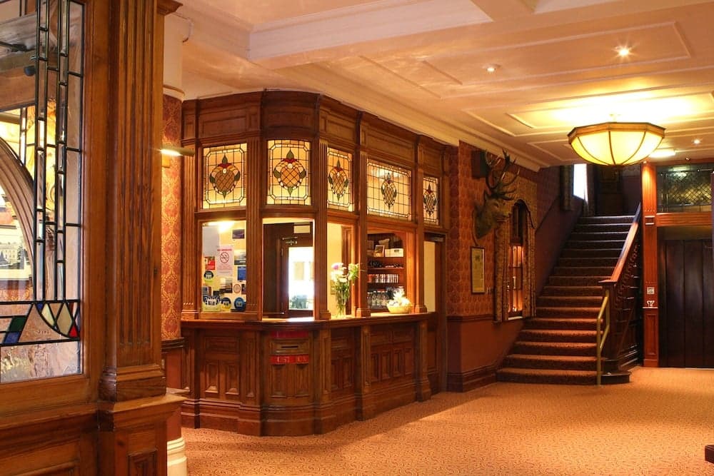 A warmly lit hotel lobby features ornate wooden paneling, stained glass windows, a grand staircase, and a reception desk, creating an inviting atmosphere ideal for travelers seeking a classic and cozy accommodation experience.