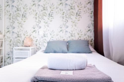 A serene hotel room features a neatly made bed adorned with a rolled towel and pillow on floral wallpaper, evoking a calm and inviting atmosphere ideal for relaxation and leisure stays.