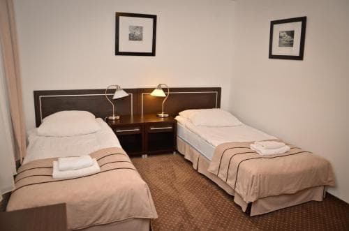 A cozy hotel room features two neatly made single beds with beige linens, a small desk with lamps, framed artwork on the walls, and a warm, inviting atmosphere ideal for travelers seeking comfort and simplicity.