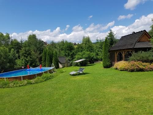 A serene backyard retreat features a vibrant blue pool surrounded by lush greenery, a cozy wooden cabin, and lounge chairs under a bright sky, ideal for a relaxing family vacation.