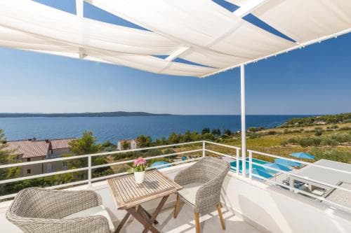 A serene balcony with wicker chairs and a wooden table overlooks the tranquil blue sea under a white canopy, ideal for a relaxing coastal getaway.