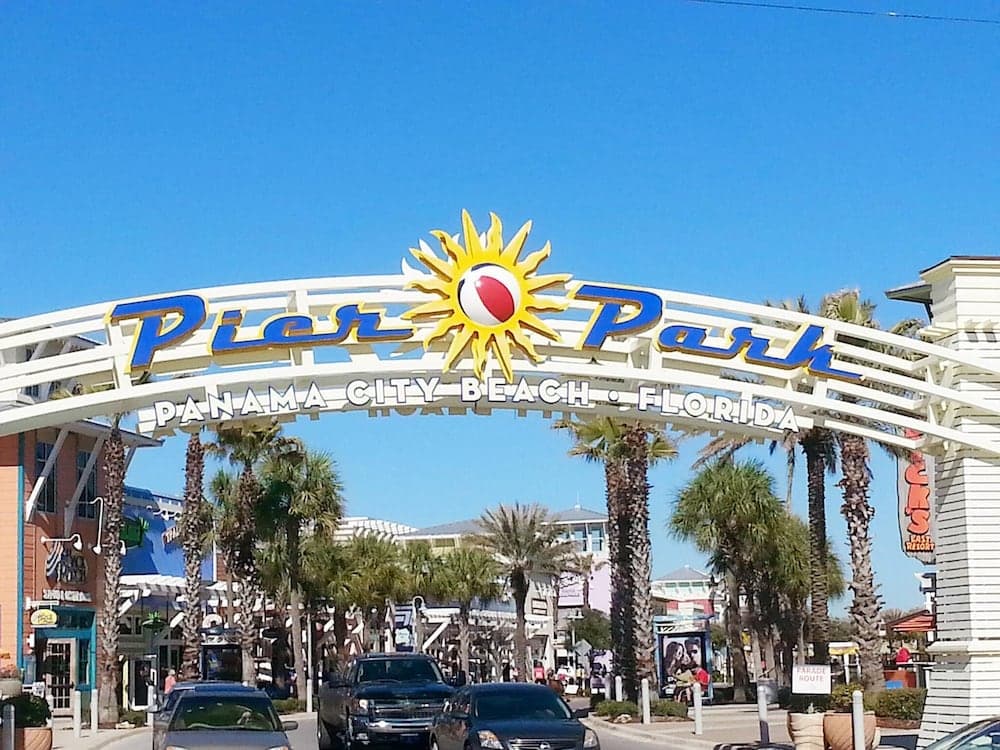 A vibrant entrance arch marked "Pier Park" welcomes visitors to Panama City Beach, Florida, under a sunny sky, framed by palm trees and bustling with cars, ideal for family vacation planning.