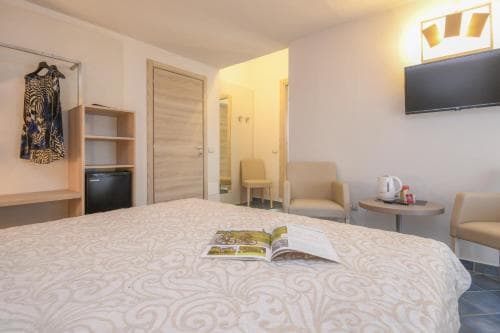 A cozy hotel room features a neatly made bed with an open magazine, a wardrobe with hanging clothes, a small seating area, and a mounted TV, creating a welcoming atmosphere ideal for travelers seeking comfort and convenience.