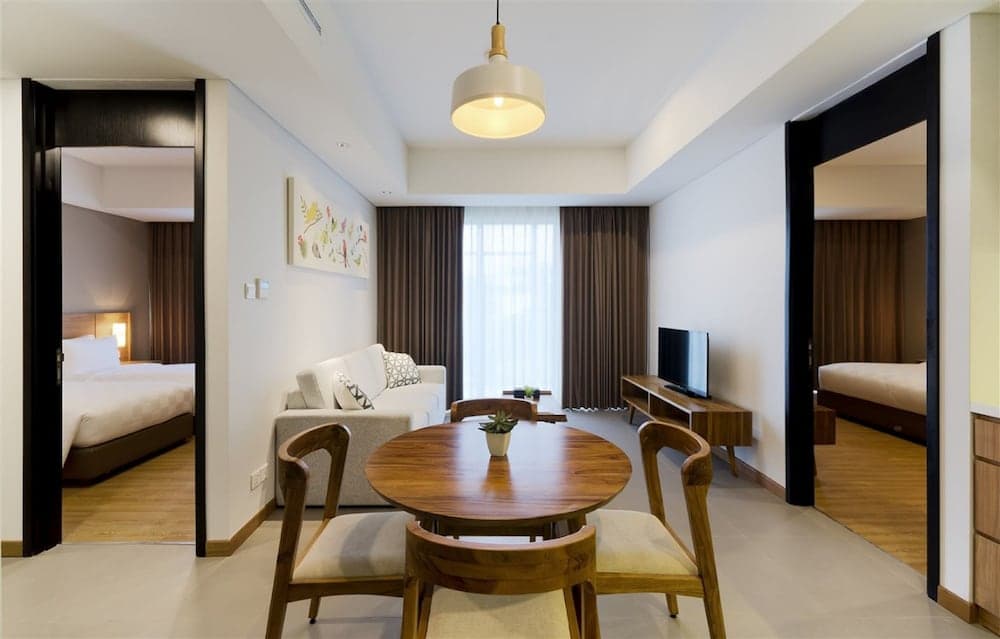 A cozy hotel suite features a modern dining area with wooden furniture, a bright living space adorned with art and curtains, and open doorways leading to bedrooms, ideal for travelers seeking comfort and style.