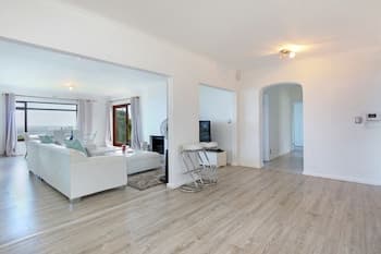 A bright, minimalist living room with white furniture, wooden flooring, and large windows overlooking the sea, ideal for a serene vacation rental experience.