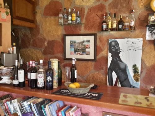 A cozy bar area with a rustic stone wall backdrop, adorned with bottles of spirits, framed art, and vibrant books, creates an inviting atmosphere perfect for travelers seeking a cultural and artistic retreat.