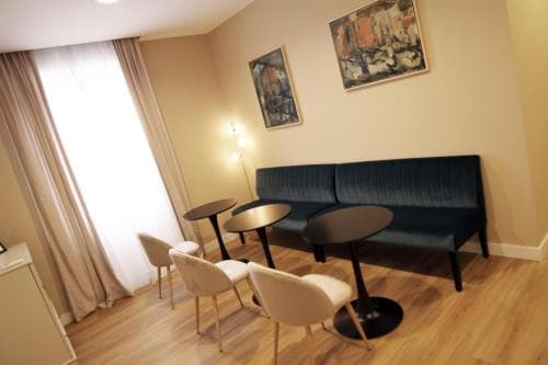 A cozy hotel lounge features modern seating with white chairs and a navy blue bench, small round tables, and warm-toned walls adorned with art, ideal for travelers seeking a quiet spot to relax or work.