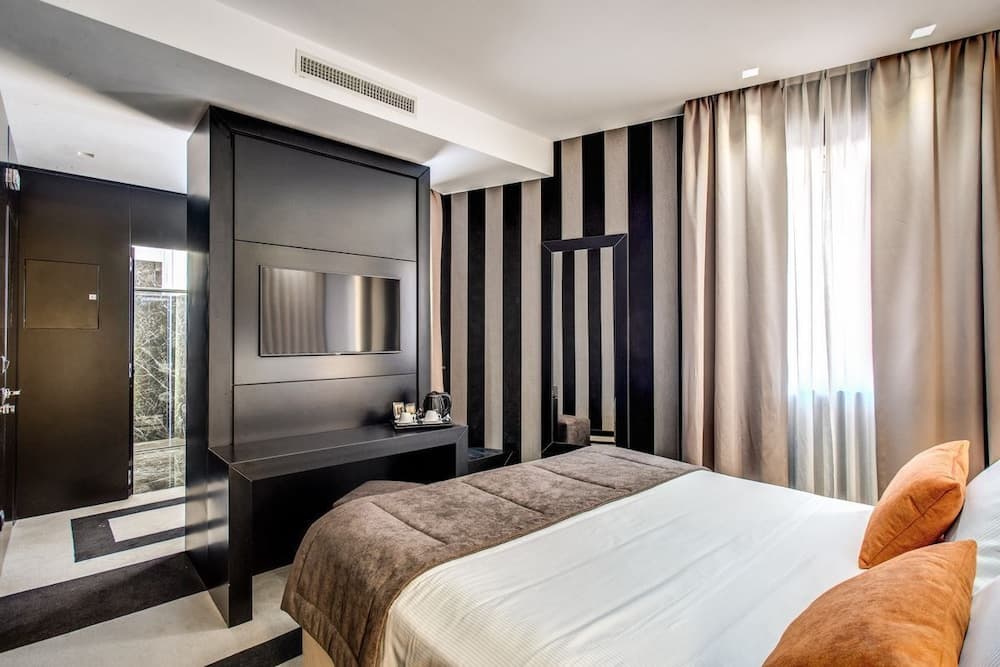 A modern hotel room features a sleek black and white striped wall, a cozy bed with orange pillows, a built-in TV console, and soft curtains, ideal for travelers seeking comfort and style.