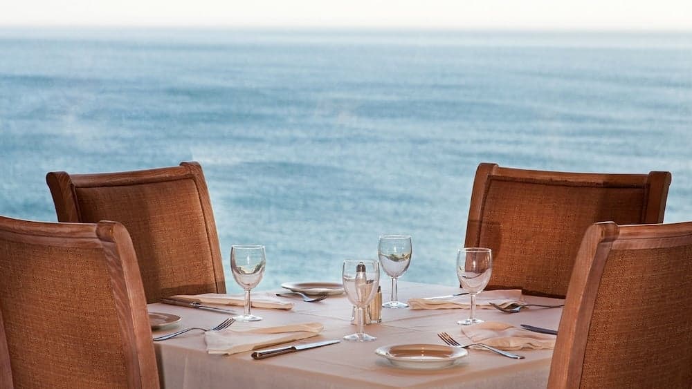 A serene seaside dining setup with wooden chairs and elegant tableware invites travelers to enjoy a luxurious meal against the tranquil backdrop of the ocean, perfect for romantic getaways or beachside relaxation.