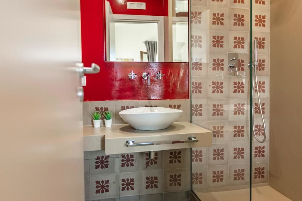 A vibrant bathroom features a red accent wall, floral-patterned tiles, a sleek white sink, and a glass shower enclosure, creating a modern and inviting space ideal for travelers seeking stylish accommodations.