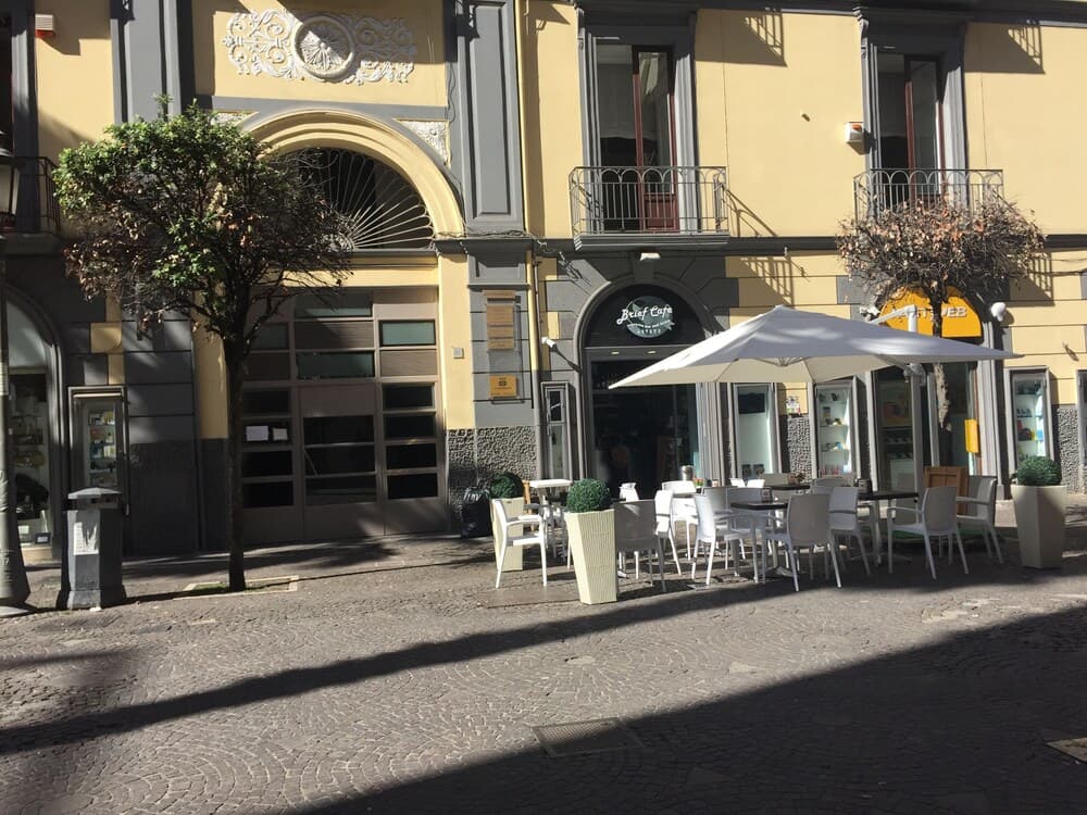 A charming European café with outdoor seating under a white umbrella invites travelers to relax on a cobblestone street lined with yellow buildings and decorative balconies.