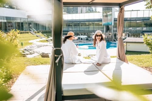 Two individuals in white robes relax on lounge chairs by a serene outdoor poolside area, surrounded by modern architecture and lush greenery, ideal for a tranquil spa retreat experience.
