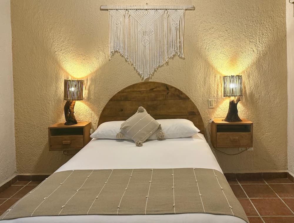 A cozy bedroom with a wooden bed adorned by a macrame wall hanging and warm-toned bedside lamps creates an inviting atmosphere ideal for travelers seeking relaxation in a boutique hotel setting.