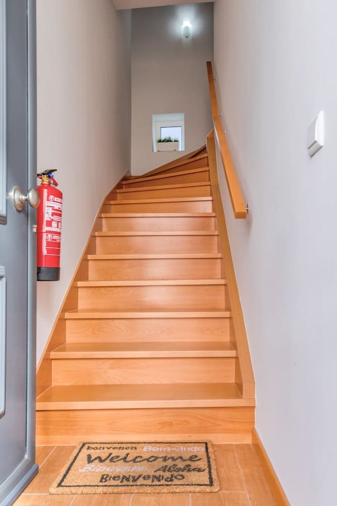 A cozy indoor staircase with wooden steps leads up to a bright landing adorned with a small window, creating a welcoming atmosphere enhanced by a multilingual "Welcome" mat at the base, ideal for travelers seeking a homey accommodation entrance.
