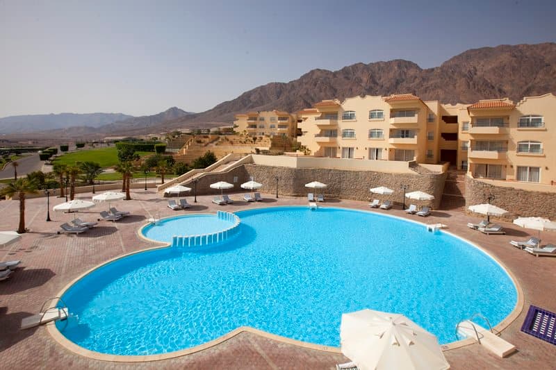 A serene resort pool area surrounded by lounge chairs and umbrellas, set against a backdrop of mountains and multi-story buildings, ideal for travelers seeking relaxation and scenic views.