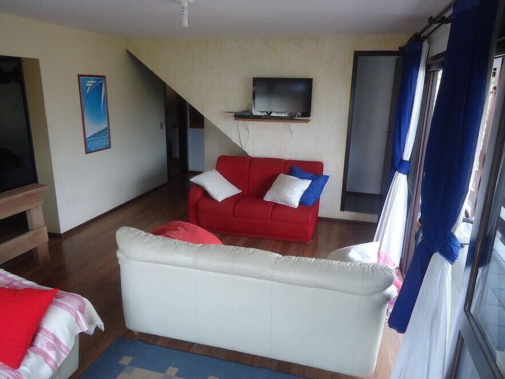 A cozy living room with a red sofa, white leather couch, wooden floors, and large windows draped with blue curtains offers a comfortable retreat for travelers seeking relaxation in a vacation rental.