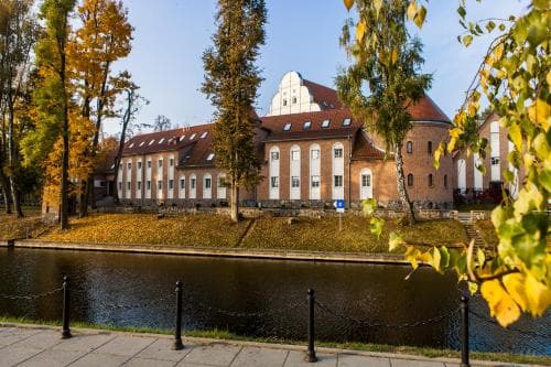 A serene autumnal riverside scene features historic brick buildings framed by golden foliage, ideal for a tranquil getaway or cultural exploration.