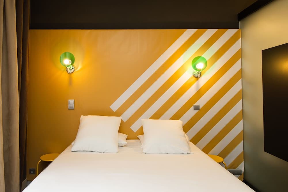 A cozy hotel room features a neatly made bed against a vibrant yellow wall with white diagonal stripes, illuminated by green wall sconces, ideal for travelers seeking a stylish and comfortable stay.