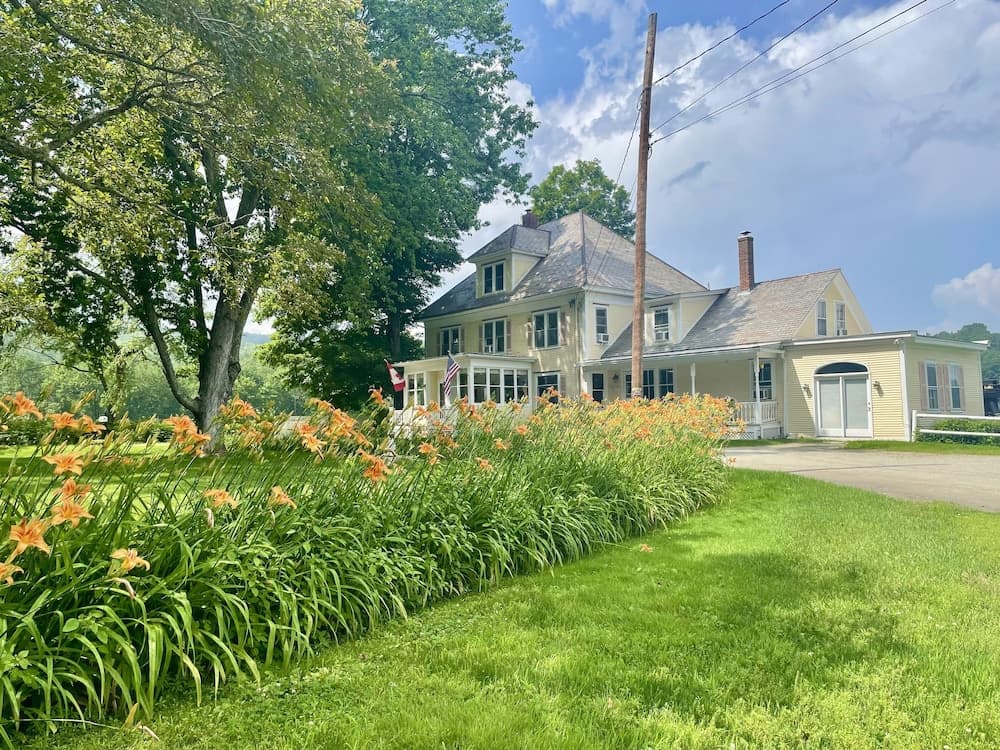 A serene countryside scene features a charming yellow house surrounded by lush greenery, vibrant orange flowers, and a large tree, ideal for travelers seeking a peaceful rural retreat.