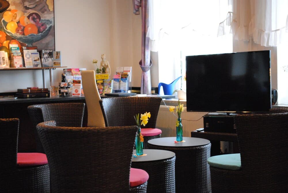 A cozy hotel lounge features wicker chairs with colorful cushions, small tables adorned with vibrant flowers, a flat-screen TV, and a bright window, creating a welcoming atmosphere ideal for travelers seeking relaxation.