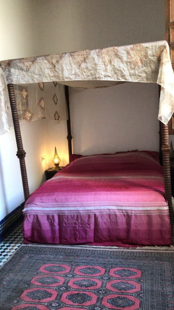 A cozy bedroom features a four-poster bed draped in patterned fabric, adorned with vibrant pink bedding, illuminated by a warm bedside lamp, set on a traditional rug, ideal for travelers seeking a charming and intimate accommodation experience.